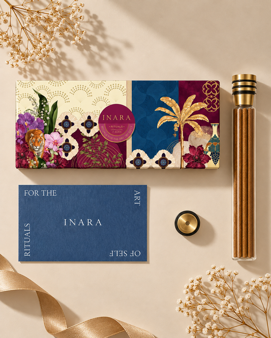 An Inara ritual set featuring a decorative illustrated box, a blue card reading 'FOR THE ART INARA RITUALS OF SELF', a gold-toned glass bottle with a gold cap, and a small round gold lid, arranged on a beige surface with dried flowers and ribbon.